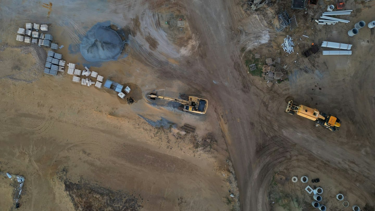 Construction vehicles and building materials sit on a sandy site.